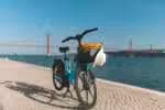 Blue electric bike with handbag and hat in basket, parked on cobblestone walkway beside Tagus river, with blurred background of suspension bridge, 25 de abril bridge